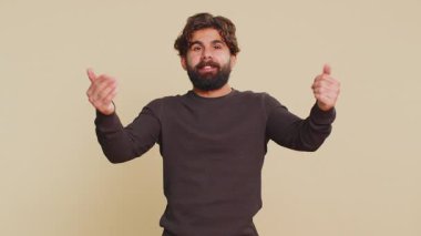 Come to me, I want to embrace you. Indian handsome man spread hands and give hug to you. Pleasant expression, love feelings. Young guy isolated alone on beige studio background. People lifestyles.