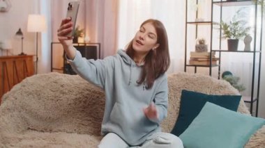 Akıllı telefondan selfie çeken gülümseyen beyaz kadın kanepeye oturmuş poz değiştiriyor ve yüz ifadeleri takınıyor. Günlük giysiler içinde mutlu çekici bir kız evde, oturma odasında vakit geçiriyor..
