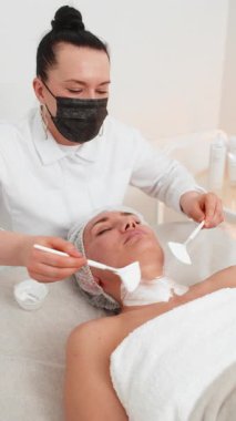 Enzim terapisi. Güzellik uzmanı, genç bir kadının yüzüne fırçayla nemlendirici krem maske takıyor. Kaplıcadaki kız cilt bakımı prosedürü. Yüz kozmetolojisi tedavisi. Dikey video görüntüsü