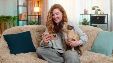 Beyaz saçlı genç bir kadın akıllı telefondan komik videolar izleyip koltukta oturmuş kahkahalarla gülüyor. Pug dog ekranı merakla izliyor. Huzurlu ortamda paylaşılan neşeli ana tepki veriyor.