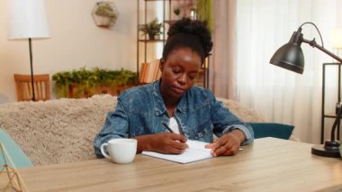 Afro-Amerikalı genç bir kadın evdeki kanepede oturmuş özenle yeni bir logo çiziyor, yaratıcı tasarım işine odaklanmış bir şekilde. Siyah kız sanatsal fikirleri ilham ile ifade eder..