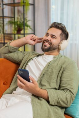 Kablosuz kulaklıklı, akıllı telefonlu mutlu genç adam evinde koltukta oturup en sevilen enerjik disko dansını dinliyor. Hafta sonu eğlence aktiviteleri.