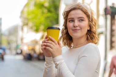 Mutlu kızıl saçlı genç kadın sabah kahvesinin tadını çıkarıyor sıcak bir içecek ve dışarıda gülümsüyor. Dinleniyorum, ara veriyorum. Şehir merkezinde dikilmek için kahve içen bir kız. Dışarıdaki kasaba yaşam tarzları.
