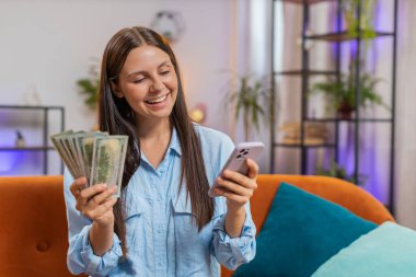 Gülümseyen mutlu beyaz kadın nakit para sayıyor, akıllı telefon kullanıyor evdeki faturaları hesaplıyor. Gelir maaşından memnun kalan ve planlı tatil ve hediyeler için para biriktiren bir kız.