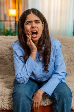 Hindistanlı genç bir kadın kanepesinde otururken aniden diş ağrısı yüzünden yanak ağrısı çekiyor. Yüzünde gergin bir ifade var. Arap kız diş rahatsızlığı ve oral sağlık sorunu yaşıyor.