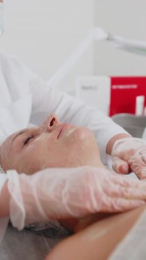 Kozmetik uzmanı, beyaz bir kadının özel nemlendirici kremini sürüyor, yüzünü ovuyor, soyuyordu. Güzellik salonunda yüz bakımı yaşlanmayı önleyen kozmetik masaj prosedürü. Güzellik tedavisi. Dikey