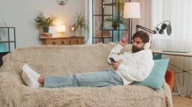 Kablosuz kulaklıklı, akıllı telefonlu mutlu genç adam evinde koltukta oturup en sevilen enerjik disko dansını dinliyor. Hafta sonu eğlence aktiviteleri.