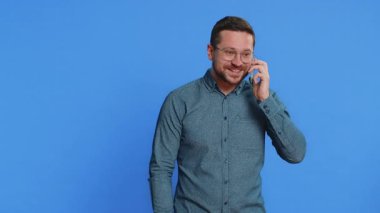 Telefon, iyi haber, dedikodu. Gözlüklü, orta yaşlı, mutlu bir beyaz adam akıllı telefondan hoş sohbetler ediyor ve arkadaşlarıyla konuşmaktan keyif alıyor. Yetişkin adam mavi arka planda kapalı alanda izole edilmiş.