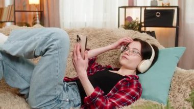 Genç kadın evdeki kanepede kablosuz kulaklık takıyor, derin bir ilgiyle sesli kitap dinlerken başını sallıyor. Kız hikayeye dikkatle ve odaklanarak tepki veriyor, ilgi çekici hikaye deneyiminden zevk alıyor..