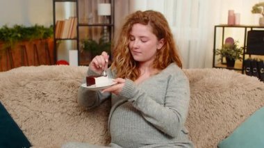 Hamile genç bir kadın, elinde bir dilim pasta tutarak kanepede oturuyor. Tatmin olmuş bir ifadeyle gülümsüyor ve tatlı tatların tadını çıkarıyor. Kız küçük zevkleri kucaklıyor, basit mutlu anlarda neşe ve huzur buluyor.