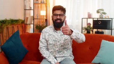 Hintli bir adam evinde oturuyor ve mutlu bir gülümsemeyle onaylama ve neşe dolu bir gülümseme sergiliyor. Arap adam rahat, modern bir dairede kanepede otururken kendini mutlu hissediyor.