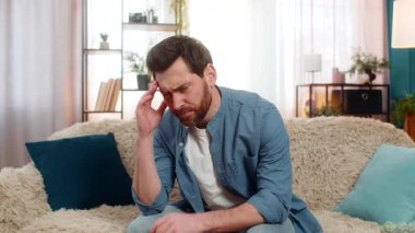 Evdeki genç adam kanepeye oturuyor, elleriyle başını tutuyor ve rahatsız bir şekilde şakaklarına masaj yapıyor. Beyaz adam şiddetli bir baş ağrısı çekiyor, stresli, yorgun ya da bunalmış, kötü bir gün geçiriyor.