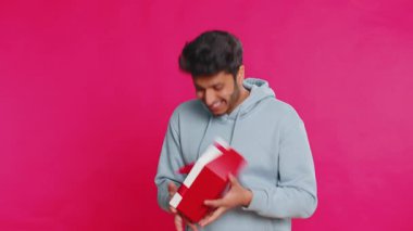 Heyecanlı Hintli adam kırmızı kurdeleli bir hediye alıyor ve bir sürprizi sevinçle kutluyor. Mutlu Arap adam pembe arka planda izole edilmiş heyecanla gülümsüyor.