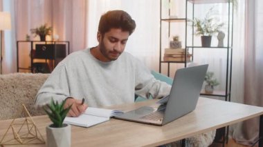 Genç Hintli adam masada oturmuş dizüstü bilgisayardaki mali raporları inceliyor, belgeleri ve makbuzları tutuyor, defterine notlar alıyor. İspanyol adam evine yakın oturuyor ve kendi işine odaklanıyor..