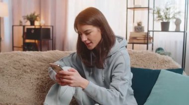 Akıllı telefon daktilosu kullanan genç bir kadın kameraya gülümseyerek tavsiye veriyor. Oldukça mutlu bir yetişkin kız. Günlük kıyafetleri içinde. Evde, oturma odasında, kanepede oturuyor..