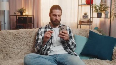 Genç adam akıllı telefonuyla evde oturuyor, mobil oyun mağlubiyetinden dolayı ani hayal kırıklığı yaşıyor. Kanepedeki adam derin derin nefes alıyor, kasvet, hayal kırıklığı, kötü haber, servet kaybı, başarısız sonuç.