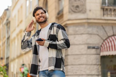 Kablosuz kulaklıklı mutlu Arap genç turist açık havada akıllı telefondan favori enerjik disko müziğini dinliyor. Şehir sokağının yanından geçen Hintli turist..