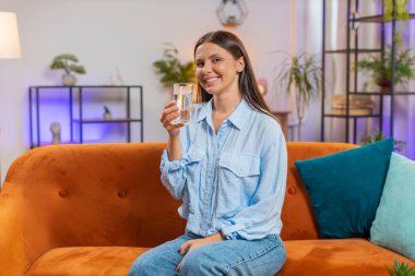 Susamış kadın elinde bir bardak doğal filtrelenmiş su tutuyor. Oturma odasında susuz kalmasın diye bir bardak su içiriyor. Ters osmoz filtresinden sonra su. İyi yaşam alışkanlığı olan bir kız sağlıklı kilo verir.