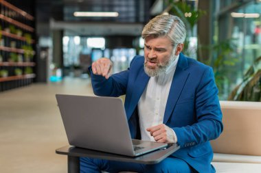 Takım elbiseli kızgın yetişkin patron, ofiste modern işyerinde otururken bilgisayarla video görüşmesi yapıyor. Tatminsiz iş adamı yöneticileri, şirketlerin sorunları hakkında astlarıyla iletişim kuruyor..