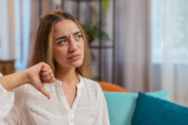 Hoşlanmamak. Mutsuz kadın portresi, hoşnutsuzluk, kötü iş, ev içinde hoşnutsuzluğu ifade eden baş parmağıyla işaret işareti gösteriyor. Kanepede oturan beyaz elbiseli kız. Yaşam biçimi