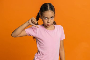 Hoşlanmamak. Üzgün, çocukluk çağındaki kız çocuğu işaret dilini göstererek hoşnutsuzluğunu, hoşnutsuzluğunu ve evde kötü çalışmasını ifade ediyor. Portakal rengi stüdyo arka planında tatsız çocuklar