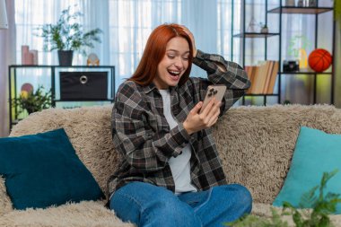 Genç ve mutlu bir kadın evde akıllı telefondan mesaj atıyor. Odaklanıp gülüyor ve kurye uygulamasında sohbet ediyor. Kanepedeki beyaz saçlı kız eğlenceli sohbetler yapıyor. Komik mesajlara sevinçle tepki veriyor.