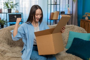 Genç, mutlu bir kadın, mutlu bir şekilde kutuları açıyor. Arzu edilen eşyaları çıkarırken hoş bir sürpriz yaşıyor. Kanepede heyecanlı beyaz bir kız, online alışveriş paketinden neşeli duygular.