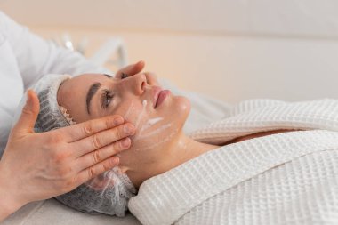 Kozmetik uzmanı beyaz kadın cilt temizleme rutini sırasında yanaklarına nazikçe köpükle masaj yaparken uzanıyor. Genç kadın klinikte profesyonel güzellik tedavisinden hoşlanıyor. Yatay