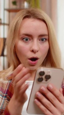 Genç yetişkin bir kadın kanepesinde oturmuş, akıllı telefondan şaşırtıcı haberler okuyor, geniş gözleri ve meraklı bakışları var. Sarışın kız internette beklenmedik bir gelişme ya da olaya hayranlıkla ve şaşkınlıkla tepki veriyor..