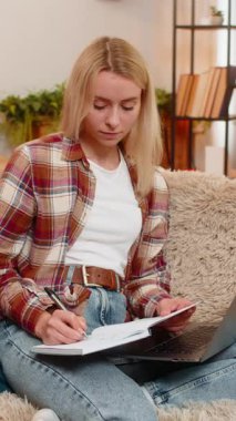 Genç yetişkin bir kadın dizüstü bilgisayar ekranına odaklanırken deftere yazar. Masrafları hesaplar. Sarışın kız dikkatli bir şekilde mali kayıtları yönetiyor, düzenli ve işine odaklı kalıyor..