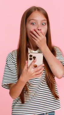 Genç kız akıllı telefondan beklenmedik haberlerle şaşırtıcı bir mesaj okuyor, gözleri açık şaşkınlıkla şaşkınlığını açıkça ifade ediyor, şaşkınlıktan nefesi kesiliyor. Pembe arka plandaki okul çocuğu.