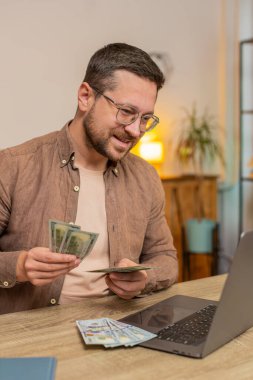 Planlama bütçesi. Zengin, mutlu bir işadamı, evdeki faturaları hesaplamak için dizüstü bilgisayarla dolar sayıyor. Kafkasyalı adam planlı tatil hediyeleri için maaş kazancından memnun..