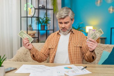 Evdeki masada oturan olgun bir adam, etrafı evrak faturalarıyla çevrili, elinde az miktarda nakit para var. Finansal zorluklar, iflas ve stres yüzünden hayal kırıklığına uğramış bir adam.