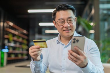 Modern ofis lobisinde online alışveriş ödemeleri için banka kartı ve akıllı telefon kullanan mutlu Çinli işadamı. İş anlaşması. Gülümseyen adam cep telefonu E-bankacılık uygulama servisiyle alışveriş yapıyor..