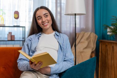 Beyaz hamile bir kadın kitap okuyor. Evdeki rahat koltukta rahatlıyor. Müstakbel anne, güzel bayan okuma tavsiyesi çocuk bakımı rehberi doğum öncesi hazırlığı. Rahatlama ve hamilelik kavramı.