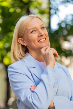 Mutlu gülümseyen, kendine güvenen, yetişkin, beyaz, profesyonel iş kadını lider resmi bir takım elbise giymiş, parmakları kenar mahalle sokaklarında dikilmiş, başarıyı düşünüyor, hayal kuruyor, gururlu, açık havada gururlu hissediyor..