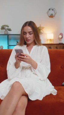 Oturma odasındaki kanepede oturan beyaz elbiseli, kafası bozuk beyaz bir kadın akıllı telefon kullanıyor. Kahverengi saçlı genç kız aniden piyango ya da kötü haberlere şaşırarak kaybediyor. Dikey.