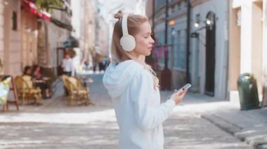 Kablosuz kulaklıkla gezen mutlu genç kadın turistin arka görüntüsü, açık havada akıllı telefondan en sevilen enerjik disko müziğini dinliyor. Güneşli şehir sokağının yanından geçen beyaz bir genç..