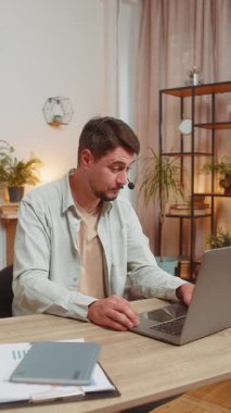 Kulaklık takan genç bir adam, serbest çalışan, çağrı merkezi ya da destek operatörü yardım hattı, müşteri ya da iş arkadaşı iletişim desteği ile konuşuyor. Ofis masasında dizüstü bilgisayar kullanan bir adam. Dikey.