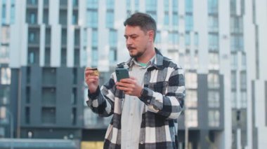 Orta yaşlı yetişkin bir adam, internet üzerinden alışveriş siparişi gönderirken kredi kartı akıllı telefonu kullanıyor. Şehir merkezinde binanın karşısında duran adam.