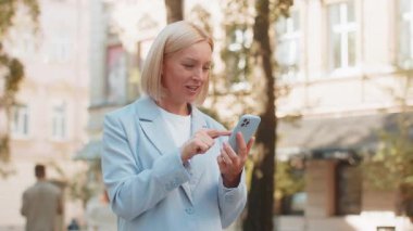 Olgun iş kadını şehir caddesinde telefonda iyi haberleri kutluyor. Beyaz, orta yaşlı, takım elbiseli işveren kadın mutluluğu ifade ediyor. Mesleki hayatta başarı ve başarı kavramı.