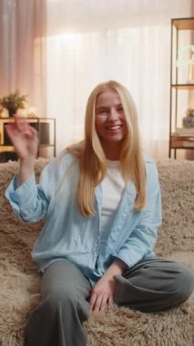 Genç bir kadın evinde oturuyor, sıcak dalgalar ve kolay iletişim için parlak bir gülümseme sunuyor. Kanepede beyaz bir kız arkadaşça el sallıyor merhaba, merhaba, günbatımı ışığı altında hoş geldin konuşması için ortam yaratıyor..