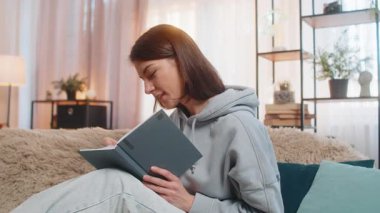 Düşünceli genç bir kadın, kalemle yazarken evdeki oturma odasındaki kanepede oturmayı düşünüyor. Gülümseyen, serbest çalışan yetişkin bir kız daire işine odaklanmış görünüyor. Çalışma Günü.