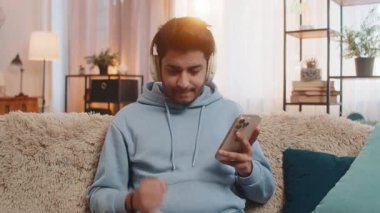 Kanepede kablosuz kulaklık takan akıllı telefondan müzik dinleyen genç Hintli adam favori pisti rahatlatmayı ve dans etmeyi seçiyor. Arap adam enerji ve neşe dolu ifadelerle ritmi seviyor..