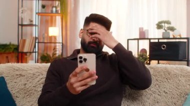 Hintli bir adam evde oturmuş hayal kırıklığıyla akıllı telefona bakıyor ve yavaşça olumsuz mesajla başını sallıyor. Kanepedeki Arap adam kötü habere üzülüyor, hayal kırıklığına uğramış ve duygusal olarak çökmüş hissediyor.