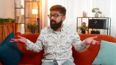 Evde oturan Hintli adam hayal kırıklığı içinde, şaşırmış ve şaşırmış görünüyor. Modern, rahat bir dairede koltukta tek başına oturan Arap adam şaşkın hissediyor.