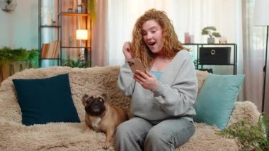 Beyaz saçlı genç bir kadın akıllı telefondan harika haberler okurken kanepesinde sevinç ve heyecanla gülümsüyor. Yanında pug dog olan bir kız var. İkisi de pozitif enerjiyle tepki veriyor..