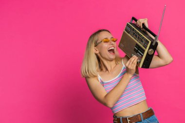 Otuz yaşında bir kadın, müzik dinlemek için retro teyp kullanıyor, disko dansı favori şarkısını dinliyor, eğleniyor, eğleniyor, klasik teknolojilerin hayranı. Pembe arka plandaki sarışın kız.