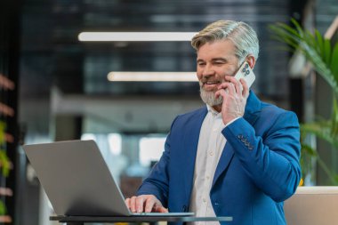 Kendine güvenen, takım elbiseli, akıllı telefon ve dizüstü bilgisayar kullanarak müşteriye cevap veren olgun bir iş adamı. Sohbet. İnternette çalışan beyaz sakallı adam modern ofis lobisinde konuşuyor..