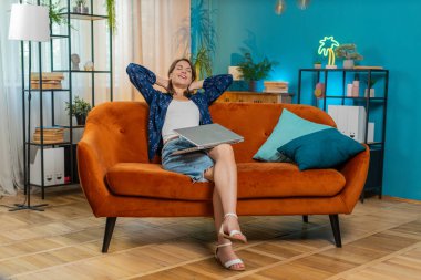 Oturma odasındaki işi bittikten sonra kanepeye oturmuş laptopu kapatan beyaz bir kadın. Mutlu genç kız serbest çalışıyor. Ev kanepesinde uzaktan kumandayla çalışıyor. Defter bilgisayarında e-öğrenme, internette gezinme